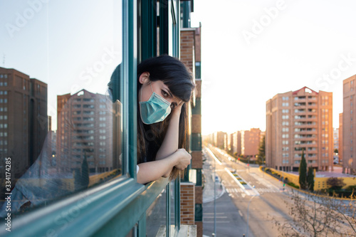 Bored woman with ffp2 face mask peeks out the window during quarantine over covid-19 crisis. Stay at home concept. Empty city under confinement. Valladolid, Spain. photo