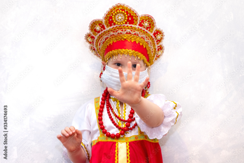 Coronavirus stop. Child 5 years old girl wearing a mask for protection and show stop hand gesture to stop the crown of the virus. National Russian clothes sundress.