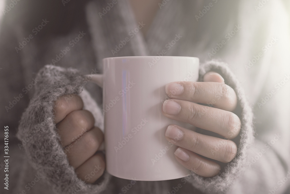 Close up of a woman hand holding a coffee mug in the morning soft sunlight. Beautiful girl in grey sweater. Close up of a woman hand holding a coffee mug in the morning soft sunlight. Beautiful girl in grey sweater.