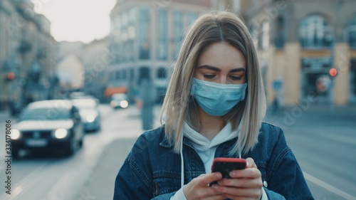 Young blonde woman in protective medical mask stands in street and uses phone texting scrolling surfs the internet search news covid19 coronavirus virus protection pandemic city close up