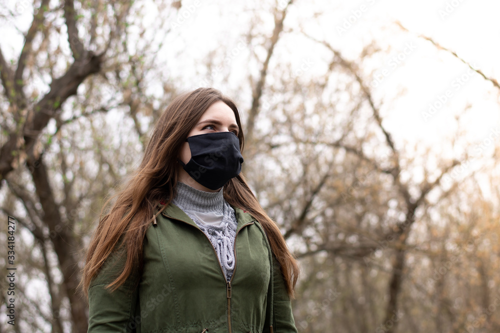 a young girl in a green jacket in a park in a black fabric mask looks away. means of protection against infections. look into the bright future