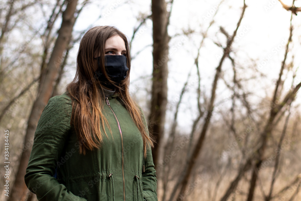girl in a green jacket in a spring park in a medical black fabric mask close-up. example of the use of protection against infections. girl in a green jacket in a spring park in a medical black fabric mask close-up. example of the use of protection against infections.
