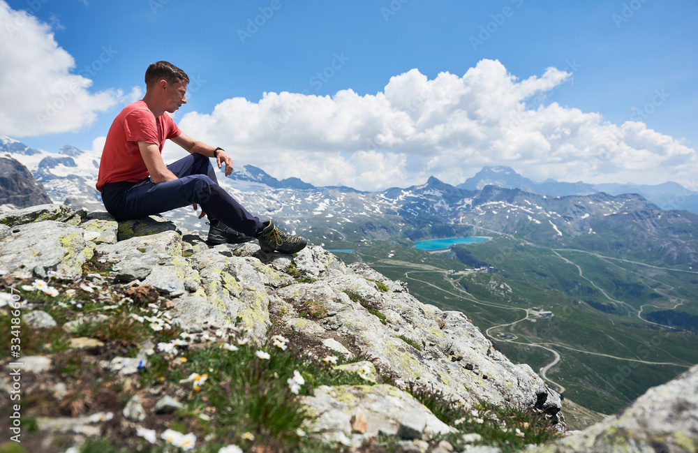Man hiker sitting on the edge of rocky hill under beautiful cloudy sky. Mountaineer admiring the view of mountain valley with grassy hills and blue lake. Concept of travelling, hiking and alpinism.