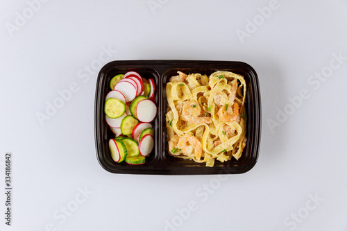 Black plastic container with pasta, shrips and fresh salad.