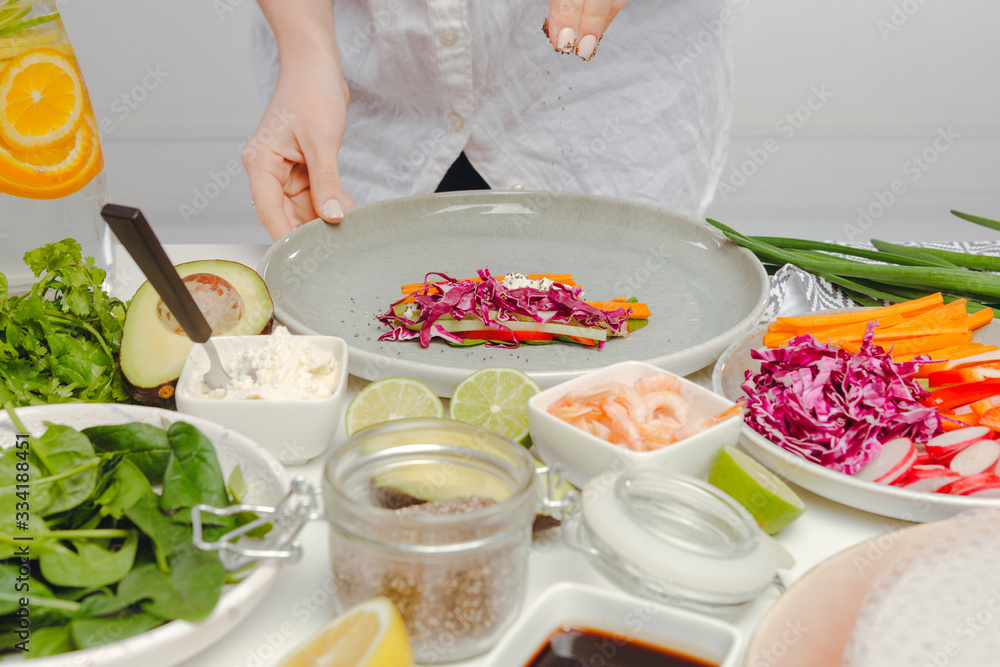 Woman's hands adding seeds to spring roll with red cabbage, cheese, carrots, cucumbers. Table with spring rolls ingredients, organic and fresh vegetables.
