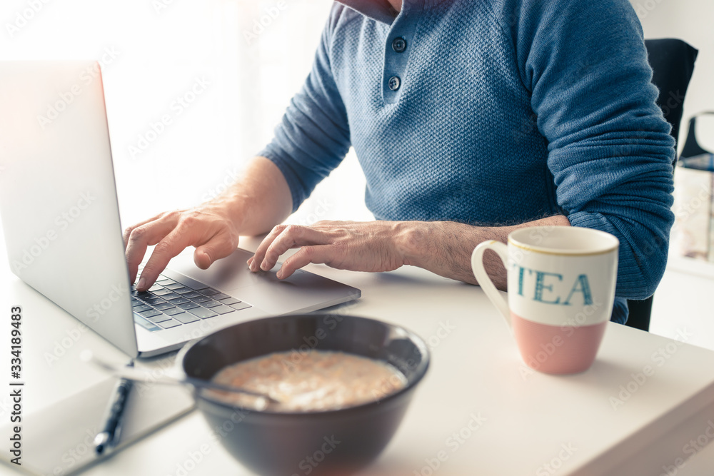 Middle-aged Caucasian man having breakfast while working from home, using his laptop, due to covid-19 coronavirus. Middle-aged Caucasian man having breakfast while working from home, using his laptop, due to covid-19 coronavirus.