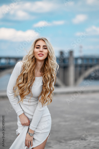 Charming long-haired blonde in a white tight-fitting dress walks around the city