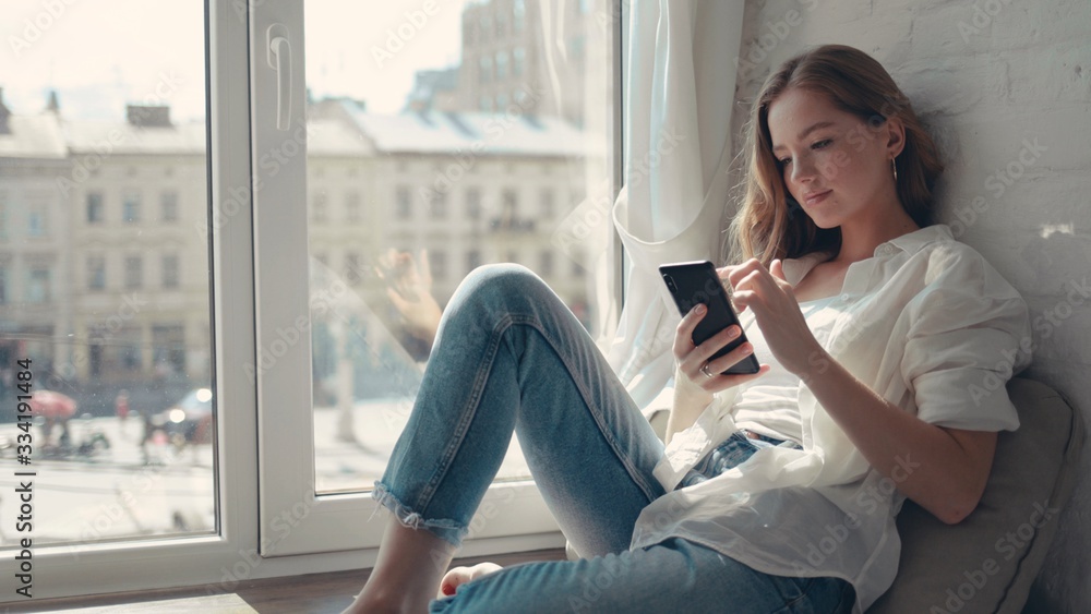 Close up pretty young woman sitting on a windowsill at home and texting on her phone communication female looking message cellphone cheerful smile use internet modern smartphone slow motion Close up pretty young woman sitting on a windowsill at home and texting on her phone communication female looking message cellphone cheerful smile use internet modern smartphone slow motion