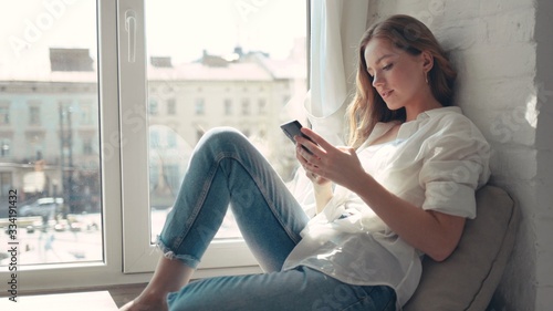 Close up beautiful young woman sitting on a windowsill at home and texting on her phone communication female looking message cellphone cheerful smile use internet modern smartphone