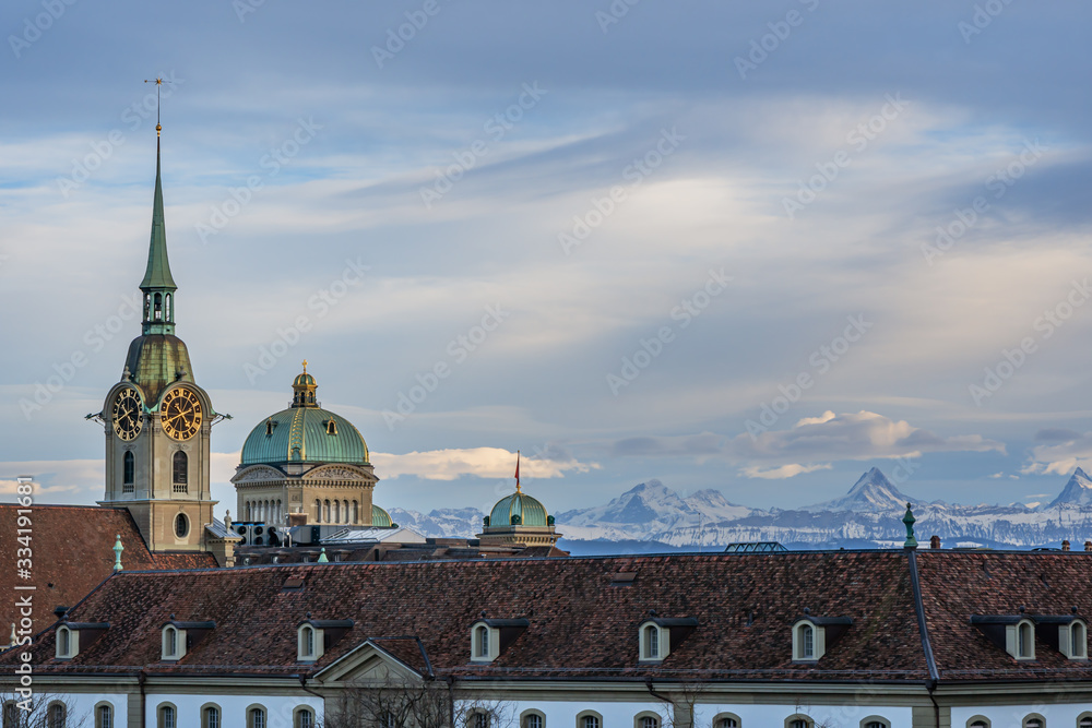 Blick auf das Bundeshaus und Heiliggeistkirche im Abendlicht vor Bergkulisse – Bern, Schweiz