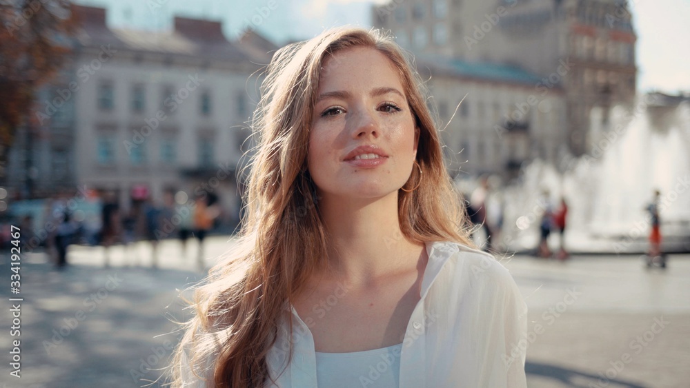 Portrait of pretty young woman with frizzy hair in city center smiling and looking at camera enjoying urban city having fun sun is shining