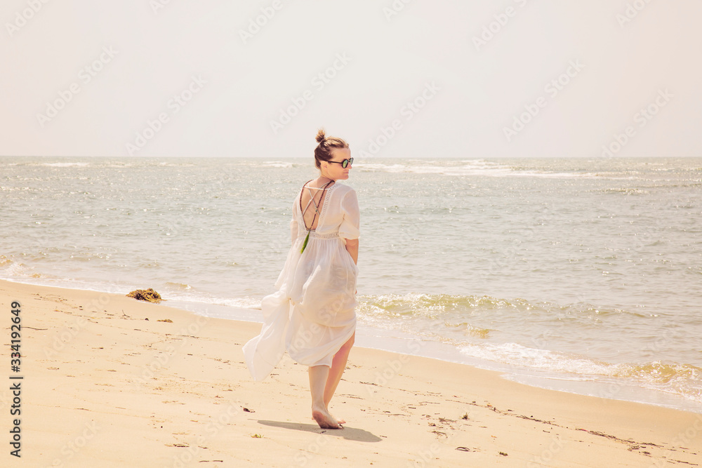 Woman in white dress and mala necklace walking away on the sunny beach by the sea Woman in white dress and mala necklace walking away on the sunny beach by the sea