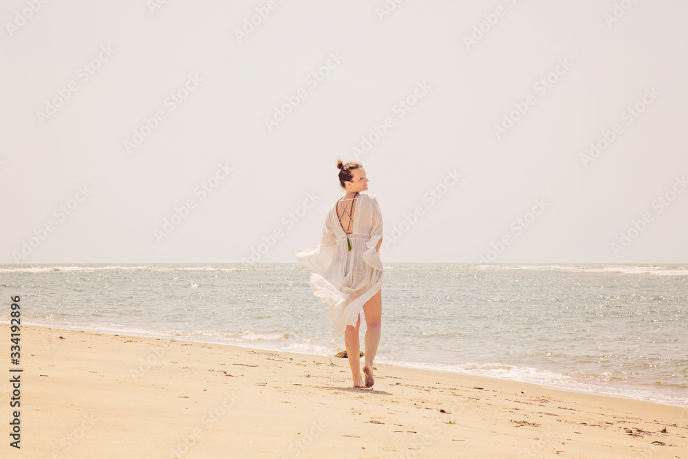 Woman in white dress and mala necklace walking away on the sunny beach by the sea Woman in white dress and mala necklace walking away on the sunny beach by the sea