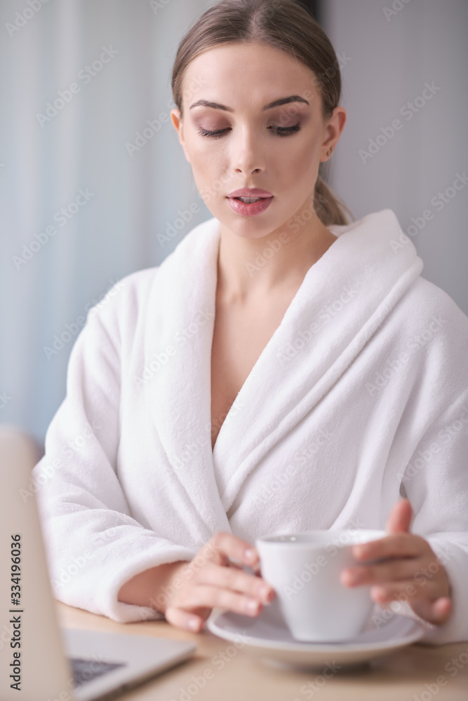 Unidentified young woman in a white bathrobe and towel sits on a cozy couch after bath surfing the Internet watches social networks using a laptop and high-speed Internet. Weekend at home concept Unidentified young woman in a white bathrobe and towel sits on a cozy couch after bath surfing the Internet watches social networks using a laptop and high-speed Internet. Weekend at home concept