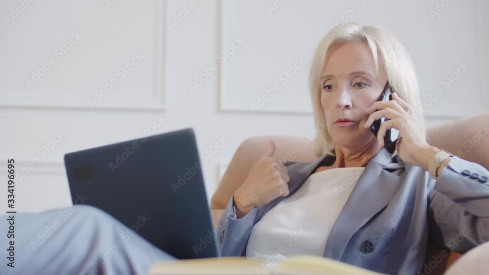 Lockdown of good-looking Caucasian woman with blond hair wearing elegant office clothes sitting on comfortable armchair in front of laptop and having cellphone chat Lockdown of good-looking Caucasian woman with blond hair wearing elegant office clothes sitting on comfortable armchair in front of laptop and having cellphone chat