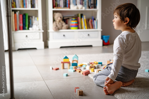 Little sad thoughtful bored toddler boy playing wooden colorful building blocks alone at home during quarantine looking at window. Child motor skills, creativity. Loneliness at self isolation period