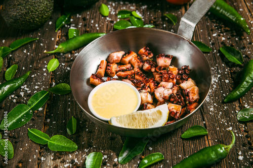 Fried octopus tentacles with oil and lemon in a metal bowl on a wooden background with decorations in the form of green leaves and green peppers © Hihitetlin