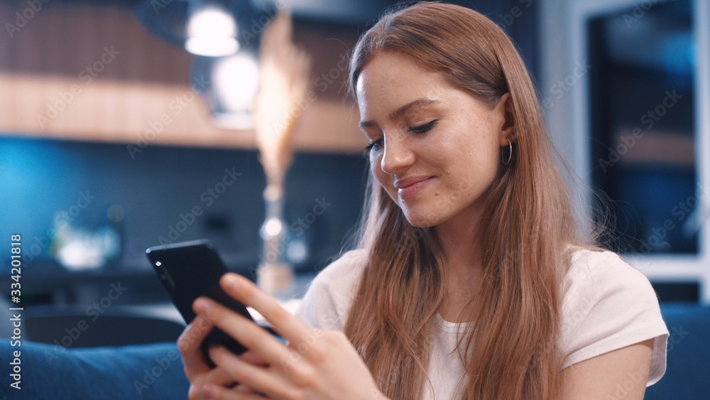 Side view of happy gorgeous young caucasian red hair woman in white t-shirt sitting on blue sofa uses phone Texting sharing messages on social media enjoy feel happy mobile technology evening relaxing