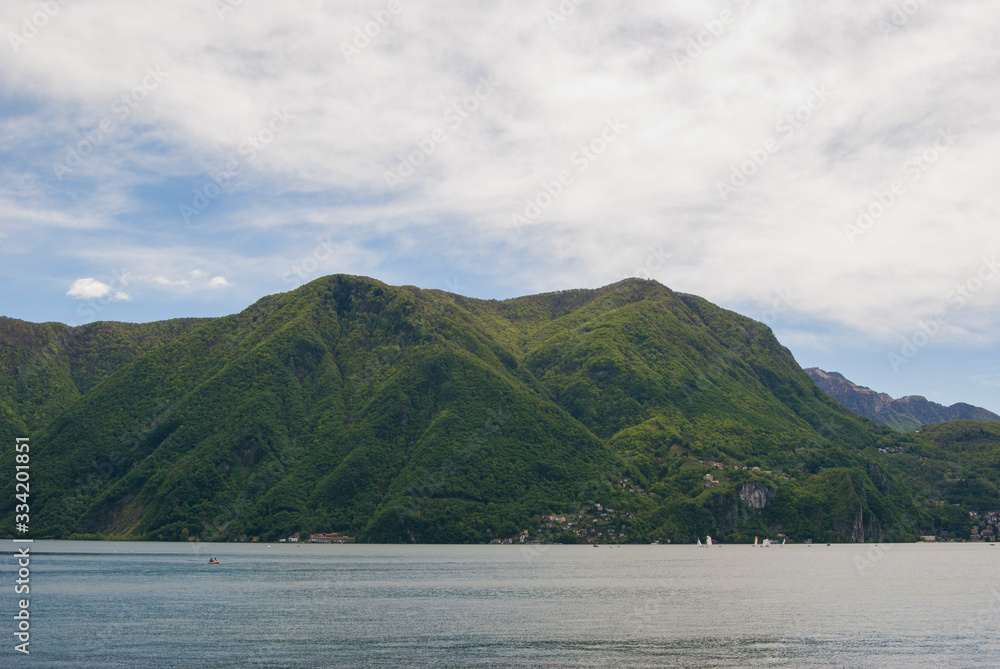 View of the lake and the alpine mountains covered with green plants in Lugano, Switzerland on a sunny summer day. 