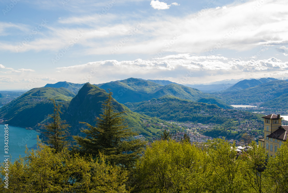 A top view of Lake Lugano, Switzerland from the height of Mount Monte Bre. Beautiful mountain scenery on a sunny summer day. View of Lake Lugano and the Alpine mountains covered with green plants.