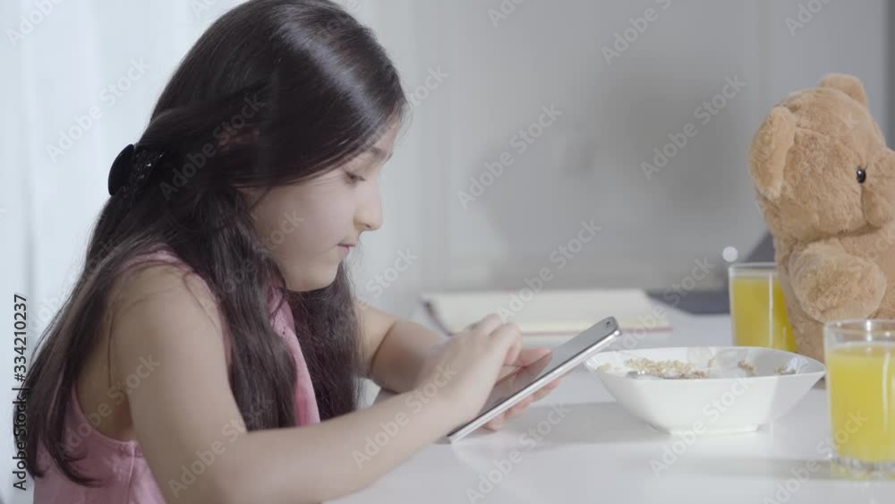Side view of concentrated little Middle Eastern girl using social media during breakfast. Cute child sitting with smartphone and meal at the table in the morning. Lifestyle, internet addiction. Side view of concentrated little Middle Eastern girl using social media during breakfast. Cute child sitting with smartphone and meal at the table in the morning. Lifestyle, internet addiction.