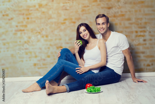 Happy smiling beautiful couple with fruits, sitting on floor, close to each other, against loft style wall. Vegetarian, weight lossing, dieting, healthy food concept photo.