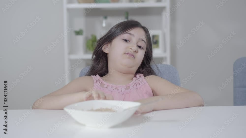 Portrait of displeased Middle Eastern girl looking with disgust at plate with porridge. Little child with black hair and brown eyes having healthy breakfast in the morning. Nutrition, childhood, diet. Portrait of displeased Middle Eastern girl looking with disgust at plate with porridge. Little child with black hair and brown eyes having healthy breakfast in the morning. Nutrition, childhood, diet.