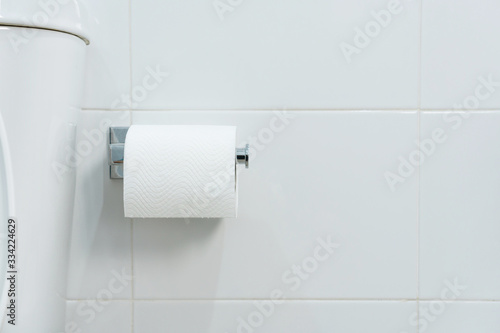 A white roll of soft toilet paper neatly hanging on chrome holder on a white bathroom wall. Close up