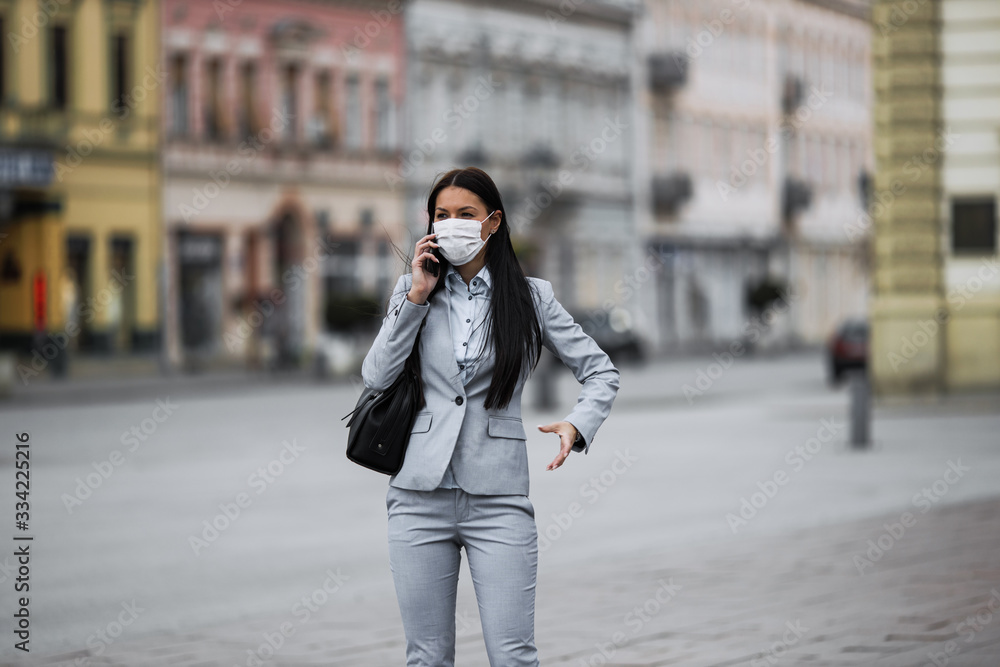Fototapeta premium Young and elegant business woman on empty city street using smart phone and wearing protective mask to protect herself from dangerous flu or virus. Corona virus or Covid-19 concept.