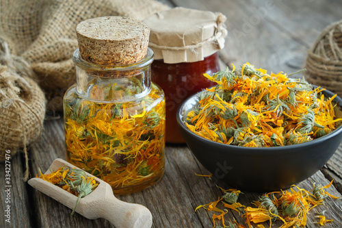 Bottles of calendula infusion or oil, healthy marigold flowers in bowl and calendula salve on wooden table. Herbal medicine.