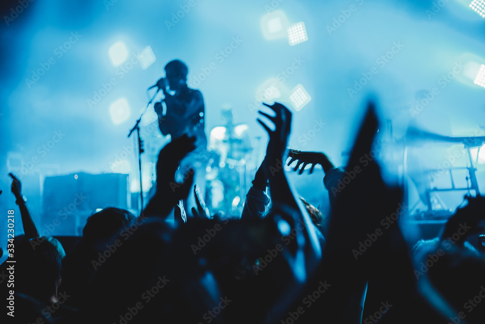 Concert crowd attending a concert, people silhouettes are visible, backlit by stage lights, raised hands.