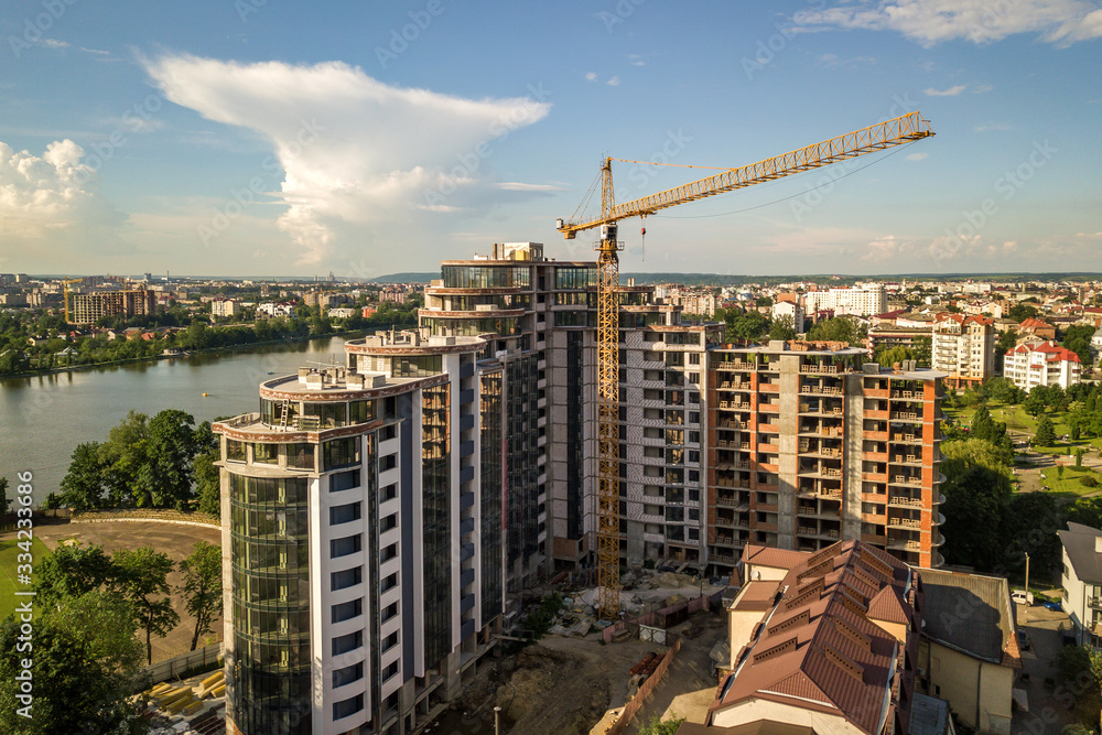 Apartment or office tall building under construction. Brick walls, glass windows, scaffolding and concrete support pillars. Tower crane on bright blue sky copy space background.