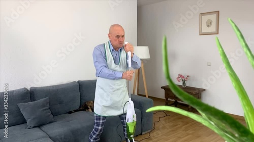 bald man at the age of 55 in home clothes and an apron is dancing happily with a vacuum cleaner, imitating playing the guitar, a cat is sitting on a sofa, a concept of household chores