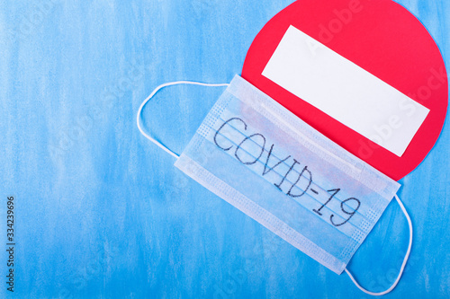 Disposable medical mask with COVID-19 written on it and no entry sign against the blue background. Restrictions caused by coronavirus outbreak 