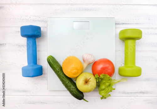 vegetables and fruits on scales, 2 dumbbells on a white wooden background