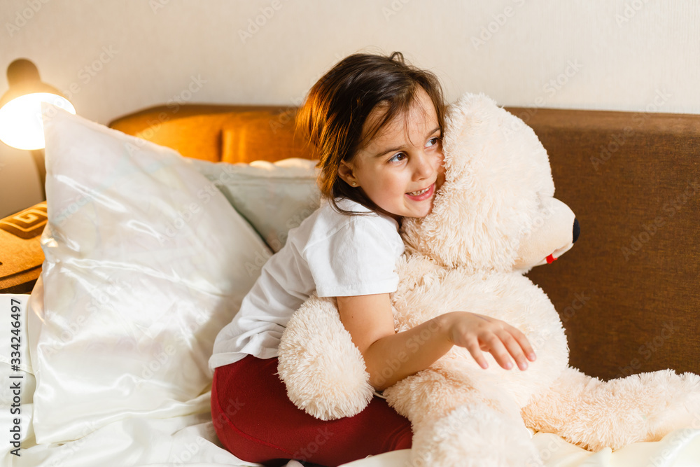 Sweet little girl is hugging a teddy bear looking at camera and smiling while sitting on her bed