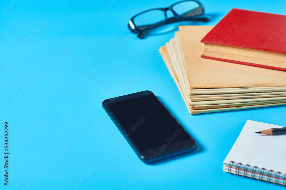 Heap of magazines, newspapers or some documents and red apple, notepad, pencil, glasses, smartphone on blue desk. Concept of education or back to school. Workplace of journalist or secretary Heap of magazines, newspapers or some documents and red apple, notepad, pencil, glasses, smartphone on blue desk. Concept of education or back to school. Workplace of journalist or secretary