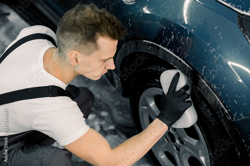 Washing a car by hand. Top angle view of young man washing the wheel of modern blue car with a sponge in carwash service