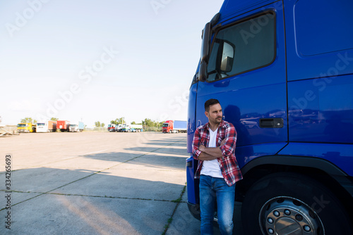 Portrait of Caucasian trucker in casual clothes standing by his truck vehicle and looking sideways.