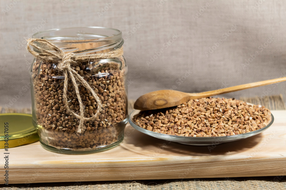 In a transparent jar and on a plate there are buckwheat grains. A wooden spoon lies on top. In a transparent jar and on a plate there are buckwheat grains. A wooden spoon lies on top.