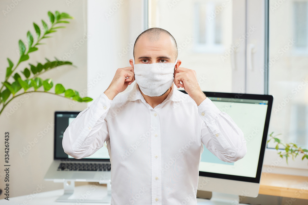 A man in a white shirt taking off a medical face mask against the coronavirus (COVID-19). An office worker at his workspace with computers and green plants in the background. Coronavirus quarantine.