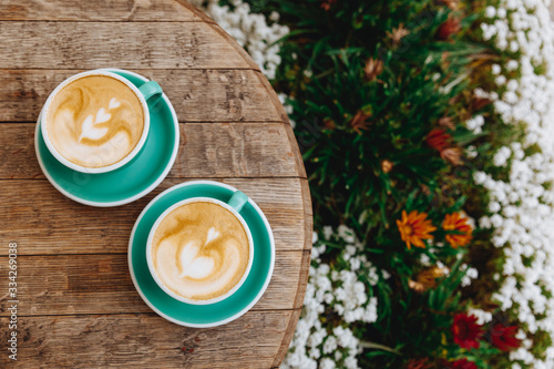 Hot aroma cappuccino with heart pattern made of milk foam in greenery ceramic mugs on round wooden table with beautiful flowers growing near outdoors. Comfortable summer terrace for coffee break