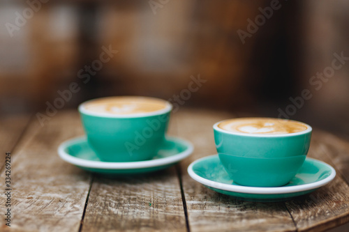 Fototapeta Naklejka Na Ścianę i Meble -  Light blue cups of hot fresh coffee with heart-shaped drawing made of milk foam on dark wooden table with blur background. Aroma drink brewed in the morning to awake