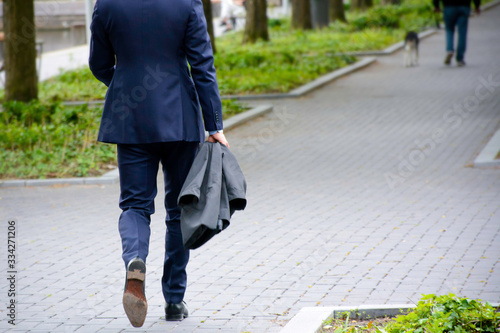 Walking businessman on sidewalk in a park. Businessman in blue business suit holding a jacket. © Evelien