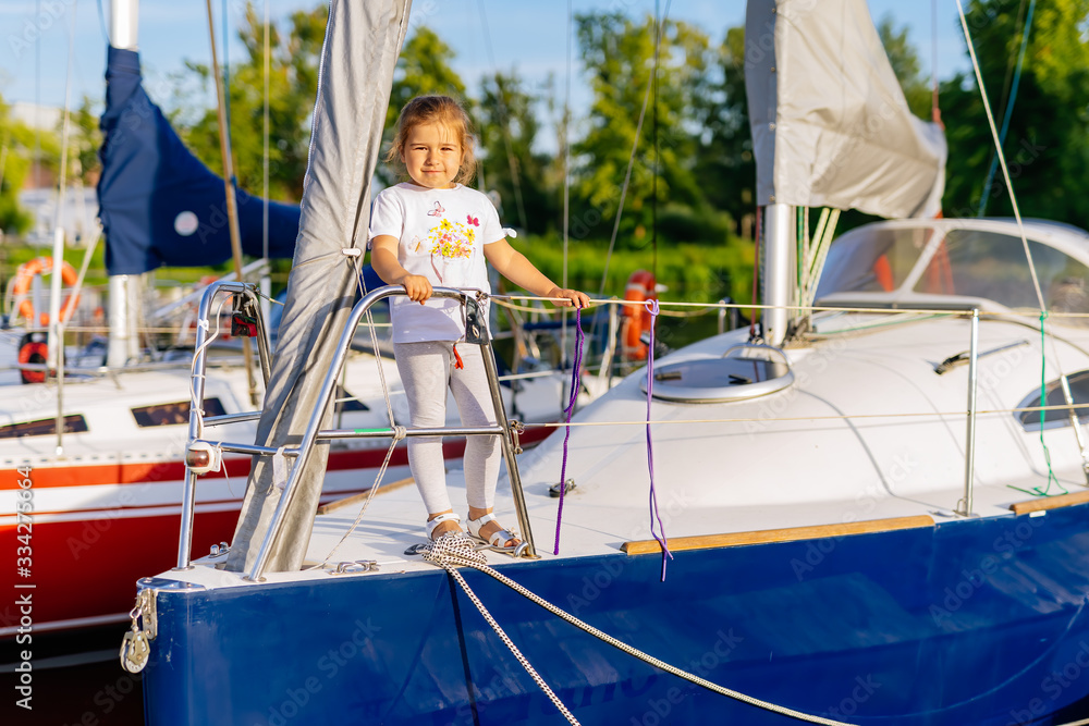 Portrait of young Caucasian white girl on prow of sailboat or yacht anchored in marina at bright sunny day. Blurred background with boats and trees