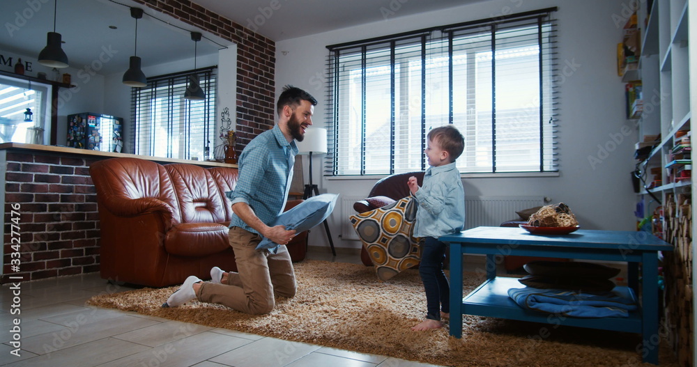 Family fun concept. Happy young European father fighting pillows with cute little son in modern light room slow motion.