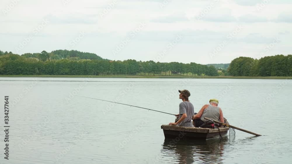Old angler with a grandson sitting in the boat on the lake with a rod while fishing. Outside. Old angler with a grandson sitting in the boat on the lake with a rod while fishing. Outside.