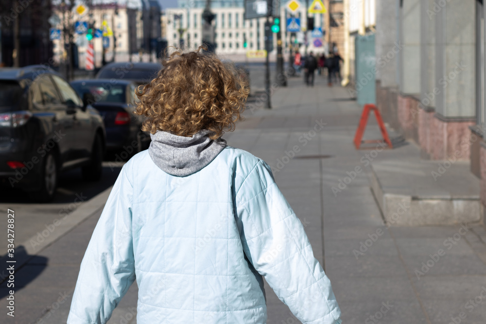 Young blonde girl with curly blond hair walks in jacket on street in sunny day. Shot from back with copy space
