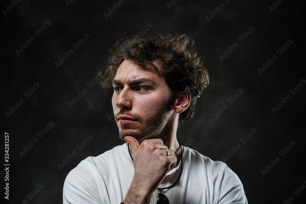 Fototapeta premium Curly and hansome caucasian man standing in a dark studio on a grey background, wearing casual white shirt looking thoughtful