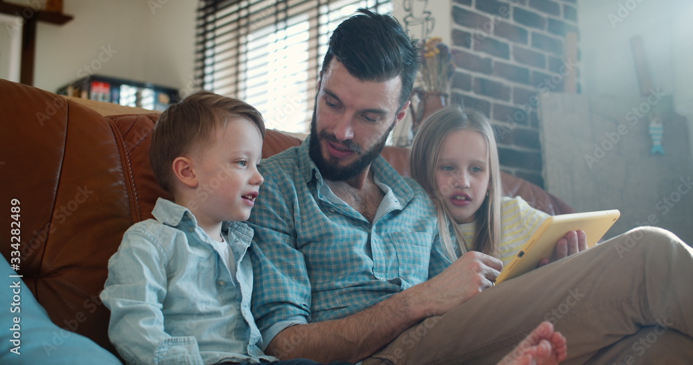Obraz premium Cute little Caucasian son and daughter read and play together with father using tablet at home on couch slow motion.